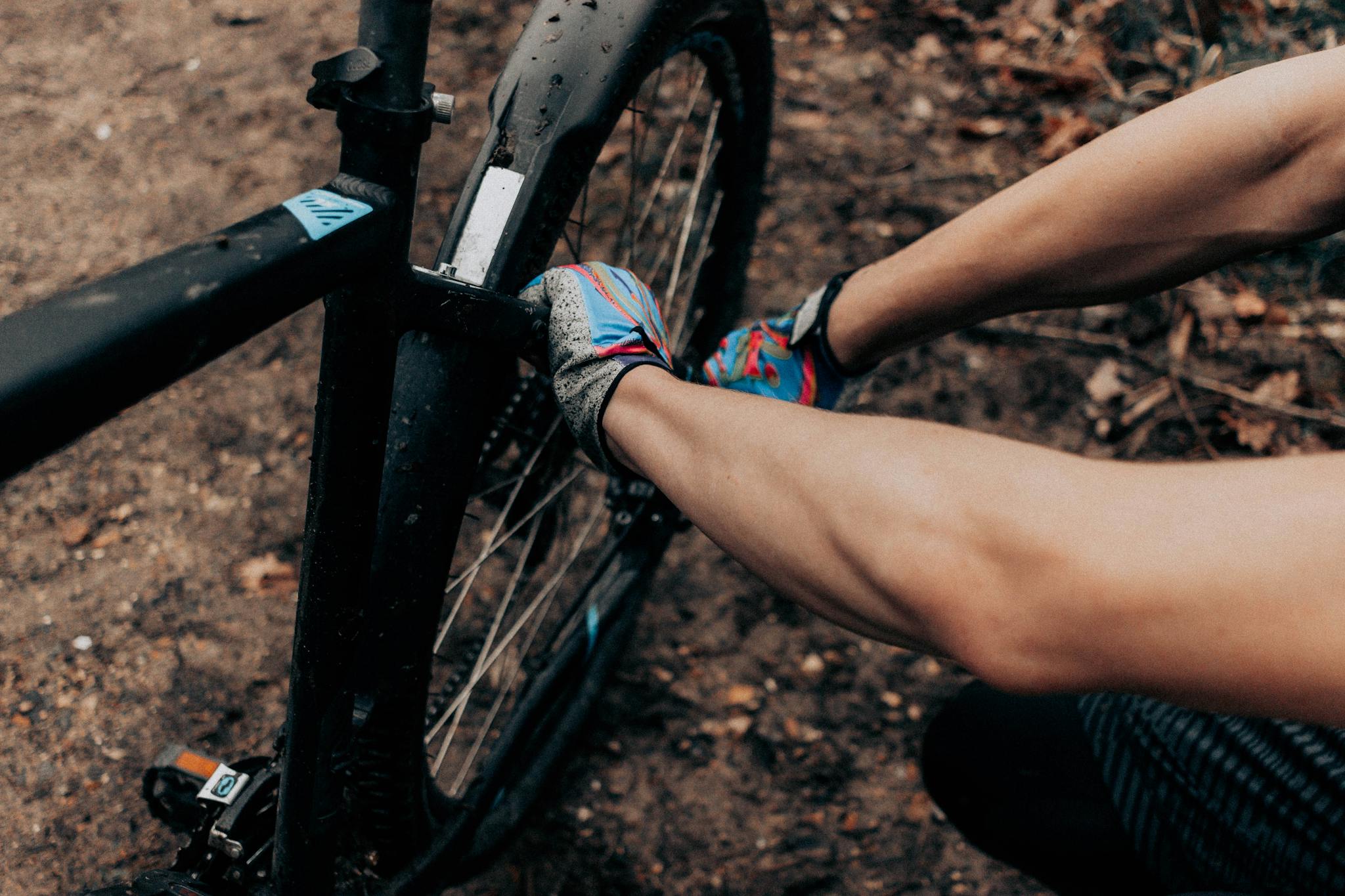 Close-up of a person repairing a bicycle on a dirt path with colorful gloves.